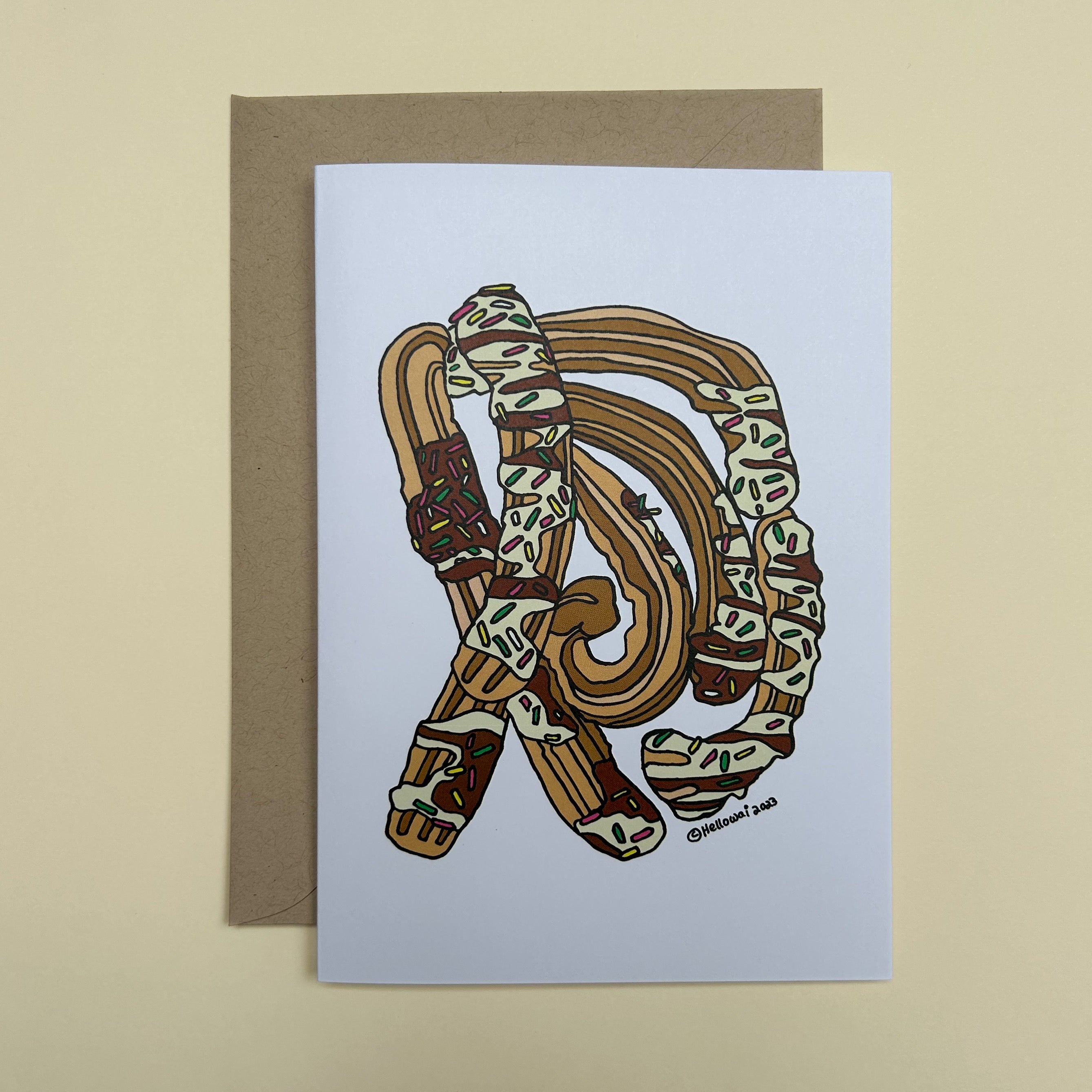 An A6 Hello Wai folded greeting card with a colour illustration of Churros covered in chocolate sauce, rainbow sprinkles, printed on white card. The card is placed on top of a brown envelope.