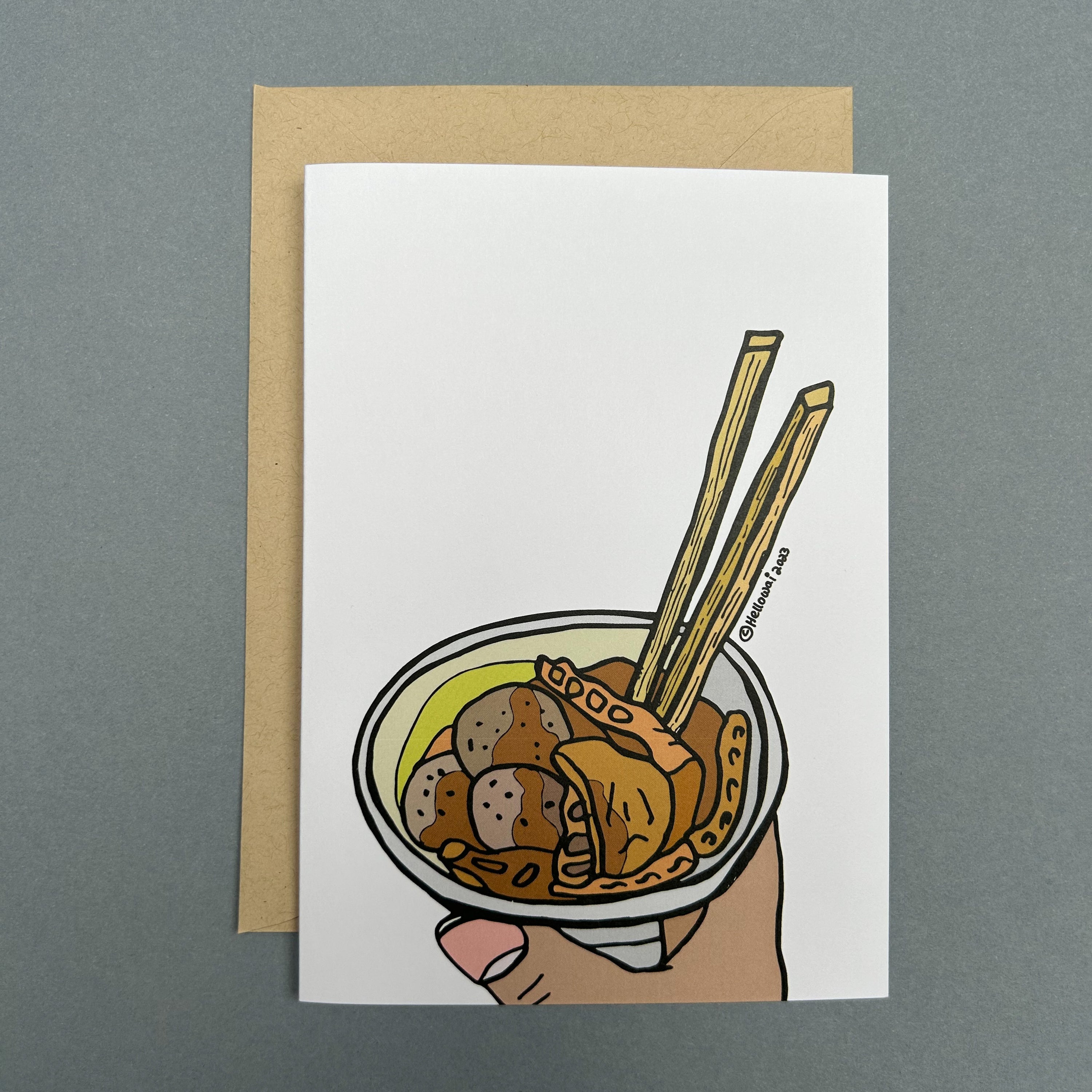 An A6 Hello Wai folded greeting card with a colour illustration of a hand holding a cup of curry fish ball and chopsticks, printed on white card. The card is placed on top of a brown envelope.