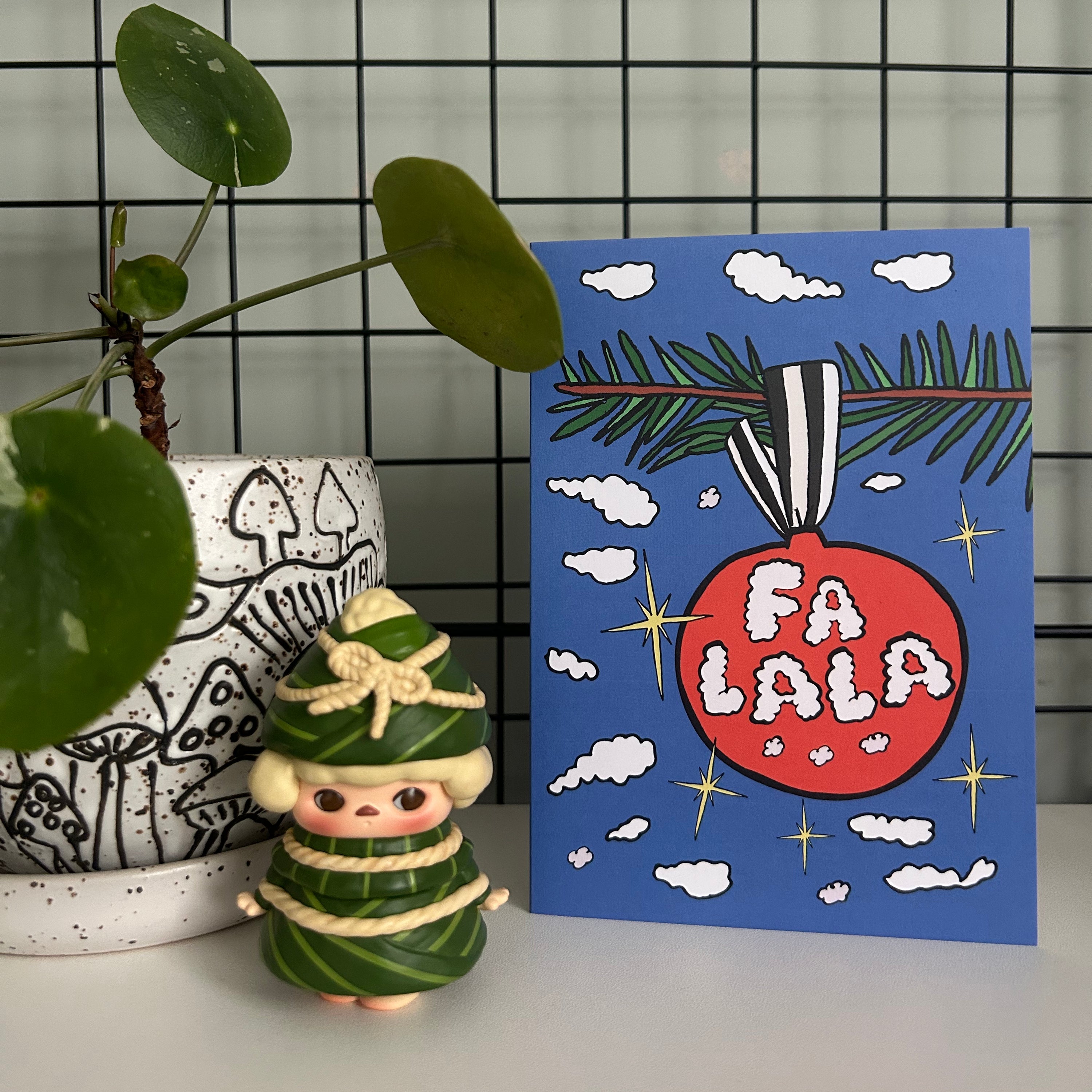 Christmas card with 'FA LA LA' design next to a decorative figure and plant.