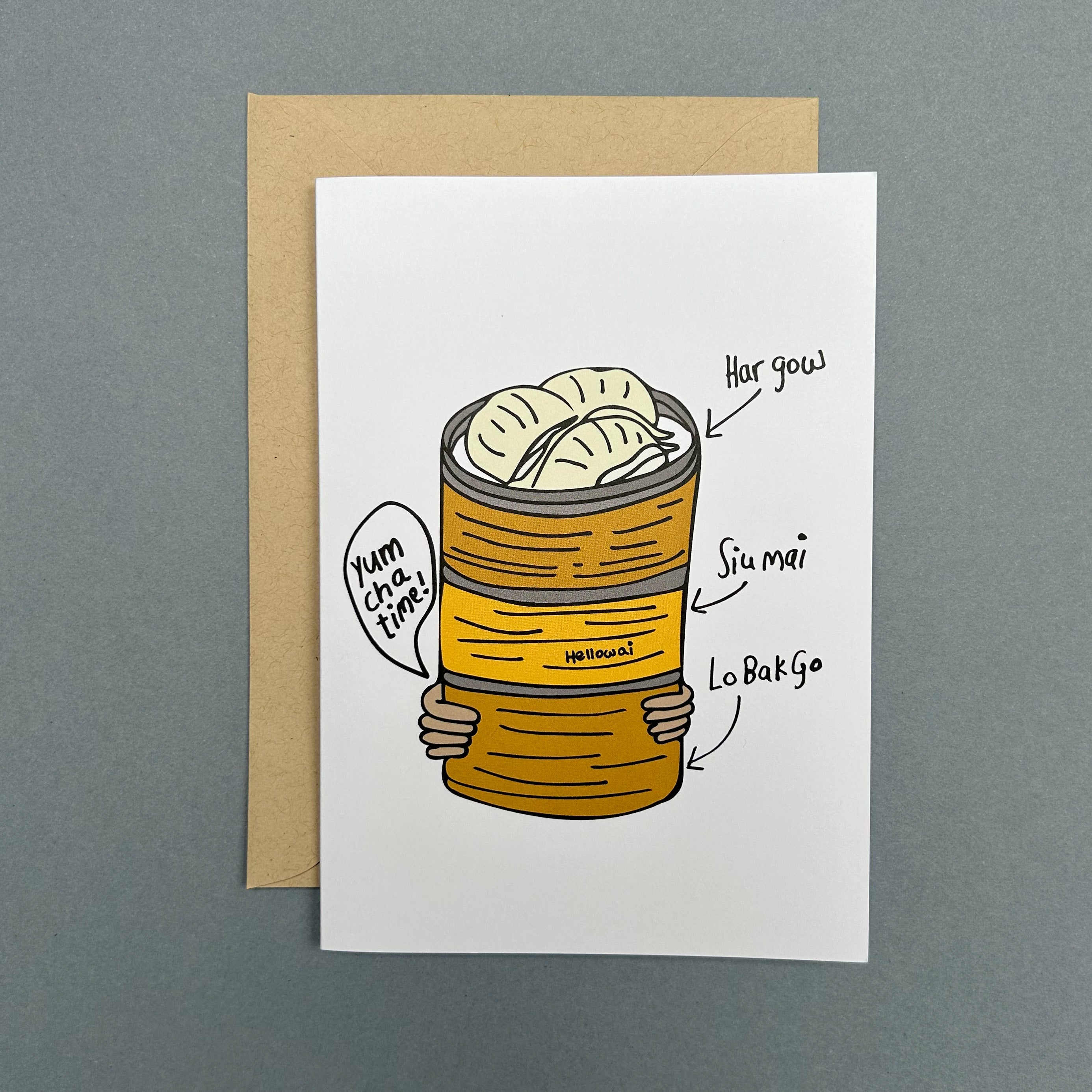 An A6 Hello Wai folded greeting card with a colour illustration of a pair of hands carrying three bamboo baskets stacked with dim sum, the top basket has har gow, printed on white card. The card is placed on top of a brown envelope.