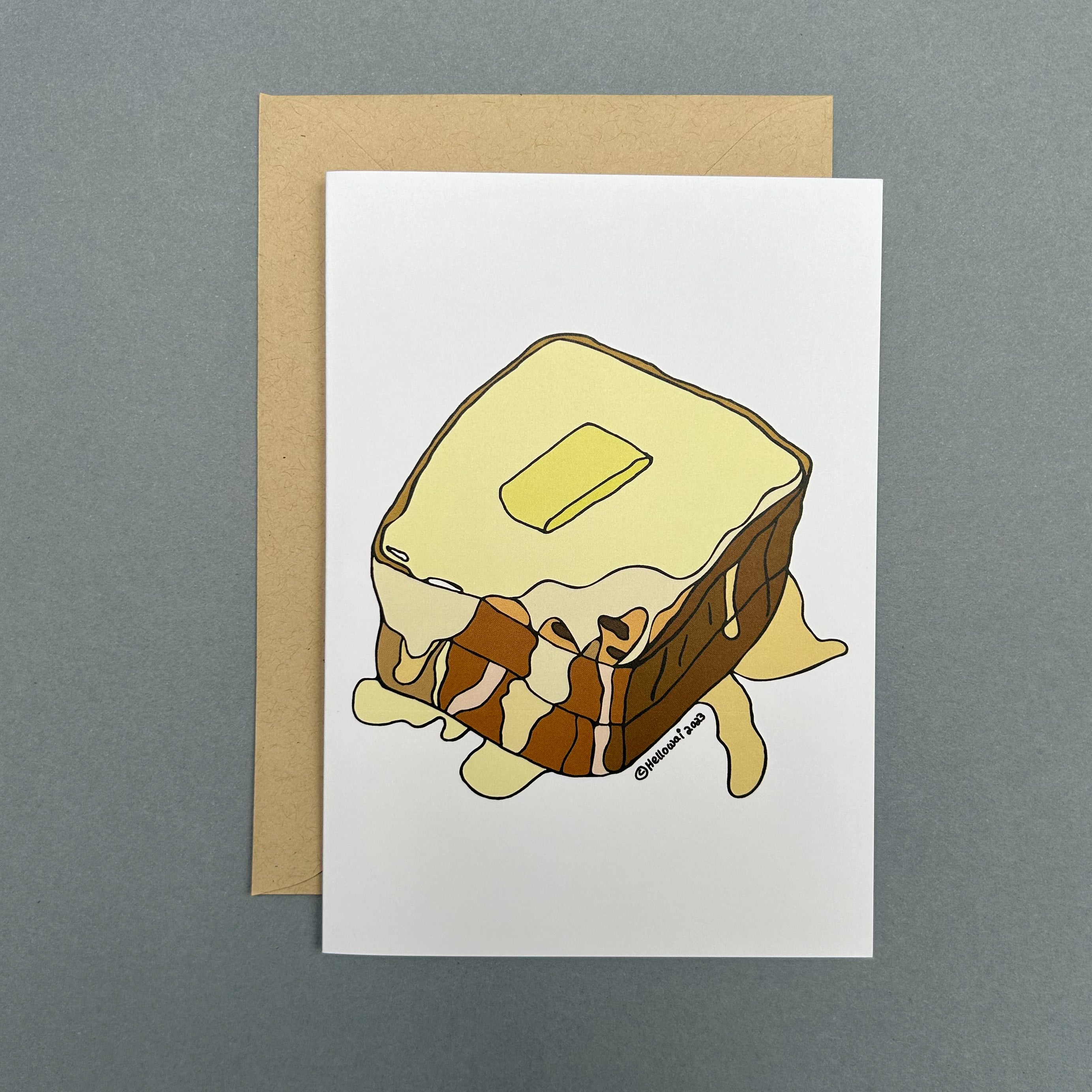 An A6 Hello Wai folded greeting card with a colour illustration of a Hong Kong Style French Toast drizzled with condensed milk and syrup and a pat of butter on top, printed on white card. The card is placed on top of a brown envelope.