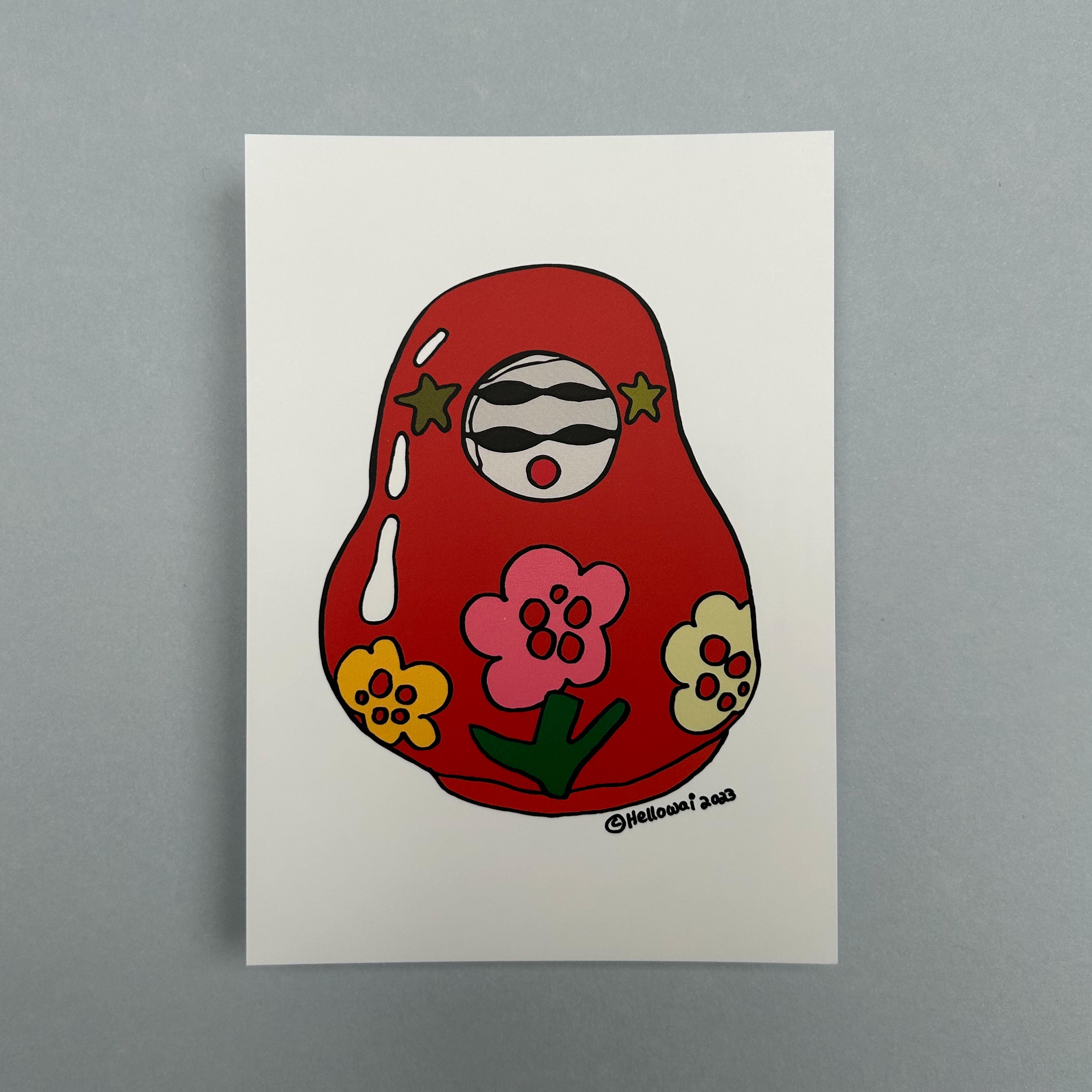 An A6 Hello Wai flat notecard with a colour illustration of a traditional Japanese roly poly doll, painted red with colourful flowers on its body, printed on white card.