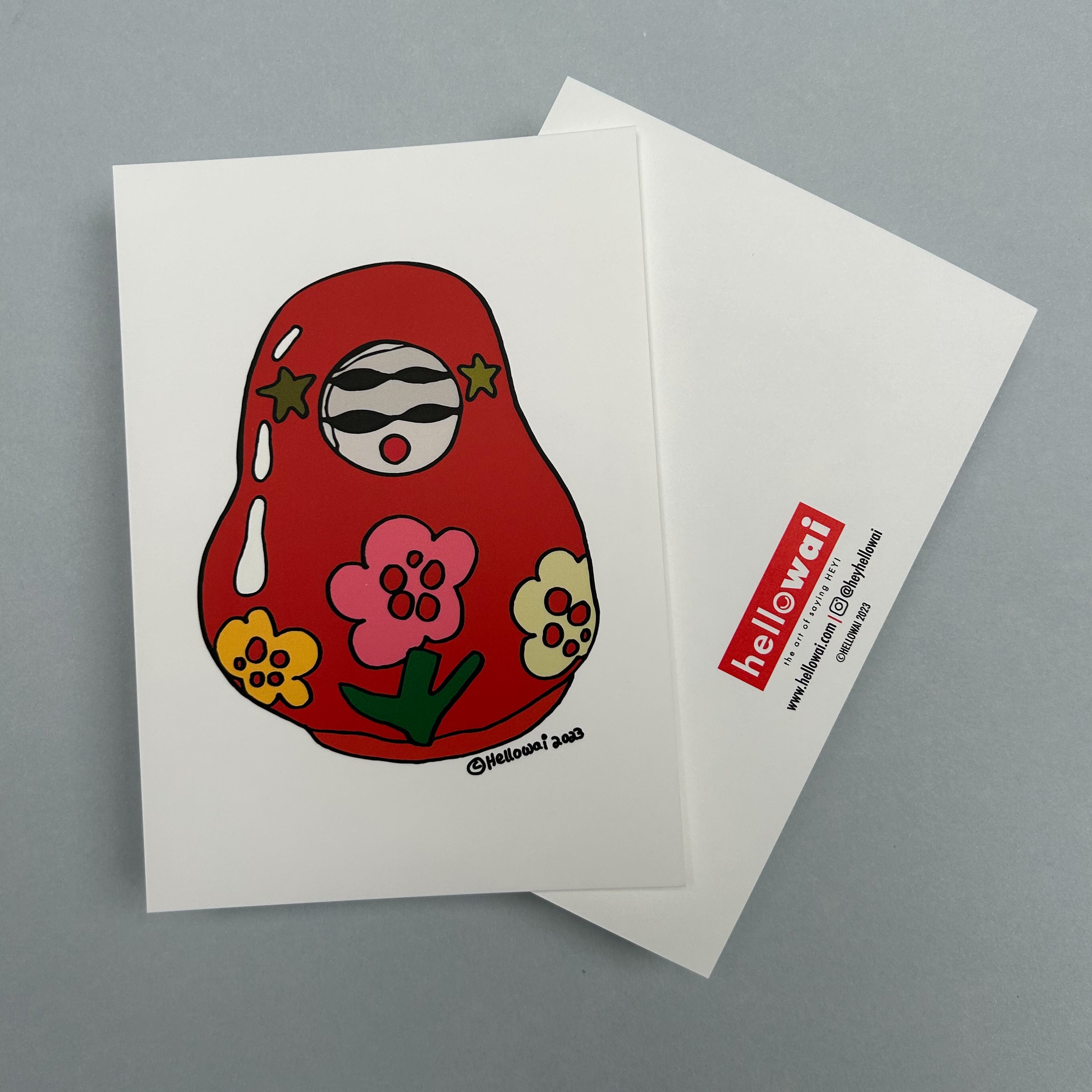 Front and back view of an A6 Hello Wai flat notecard. Front has colour illustration of a traditional Japanese roly poly doll, painted red with colourful flowers on its body. Back has the Hello Wai logo.
