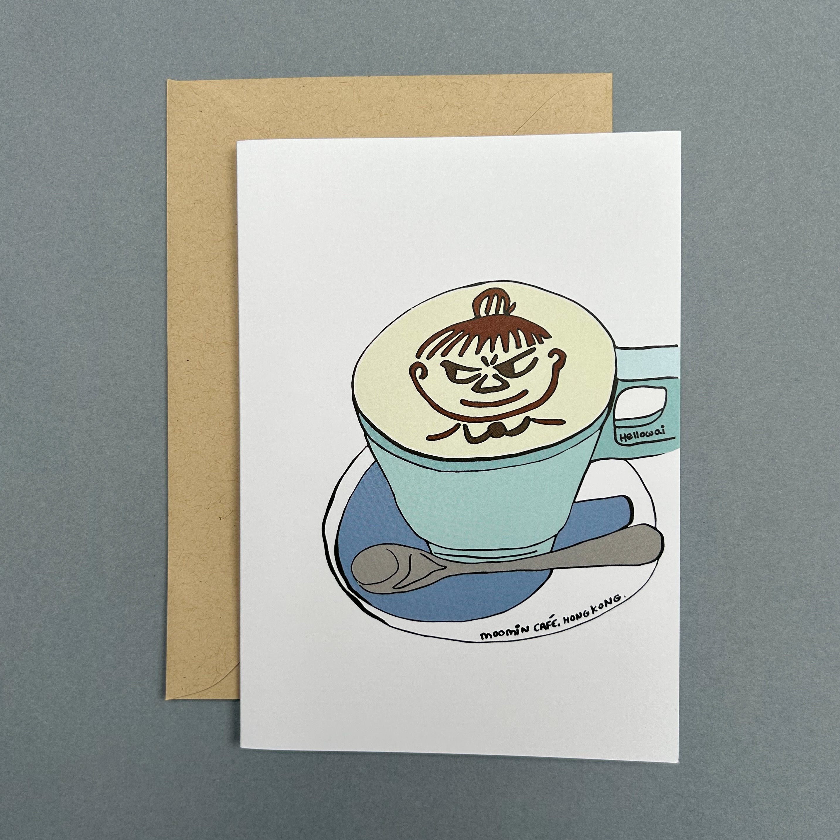 An A6 Hello Wai folded greeting card with a colour illustration of a caffe latte with chocolate cartoon character stencilled on top served in a light blue cup on a saucer with a teaspoon, printed on white card. The card is placed on top of a brown envelope.