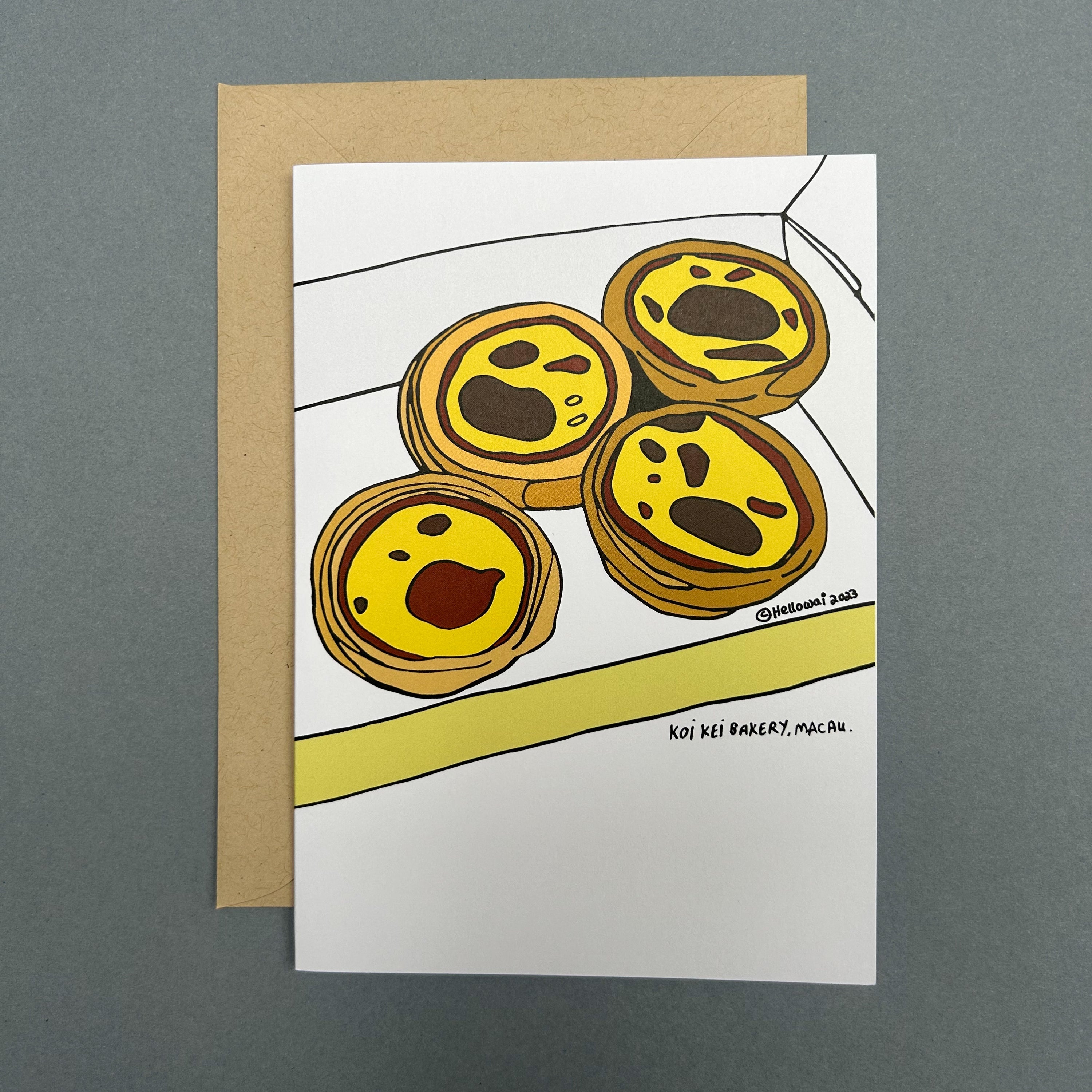 An A6 Hello Wai folded greeting card with a colour illustration of four Portuguese egg tarts in an open box, printed on white card. The card is placed on top of a brown envelope.