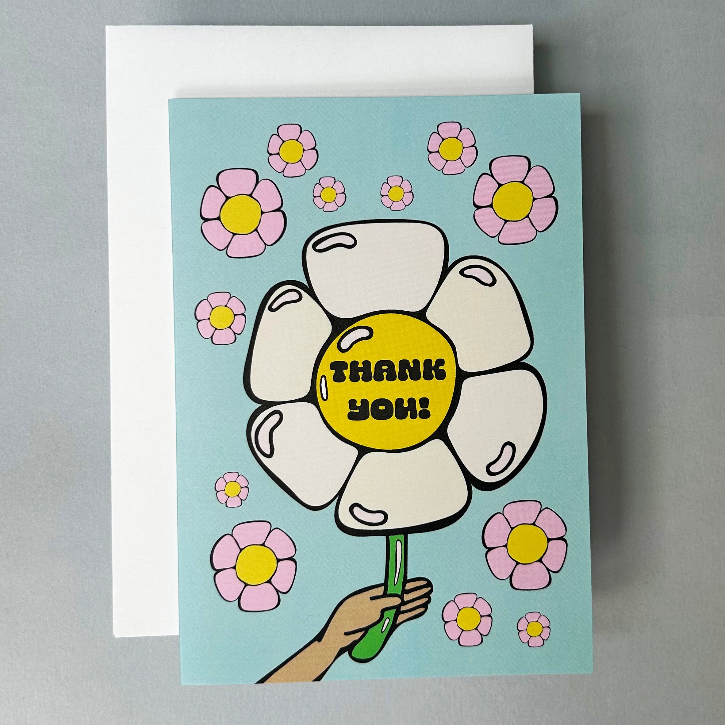 Greeting card with an illustration of a hand holding a large daisy balloon with "Thank You!' written on it. There are smaller pink daisies in the background and the card is set against a white envelope on a light grey background.