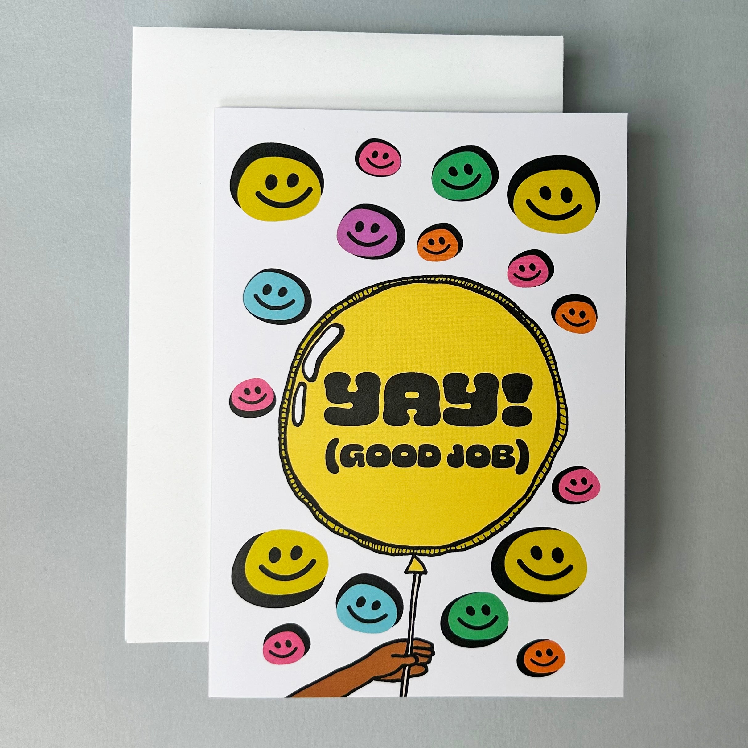 Greeting Card with illustration of colorful smiley faces and a yellow balloon saying 'Yay! (Good Job)' sitting on top of a white envelope against a light grey beackground.